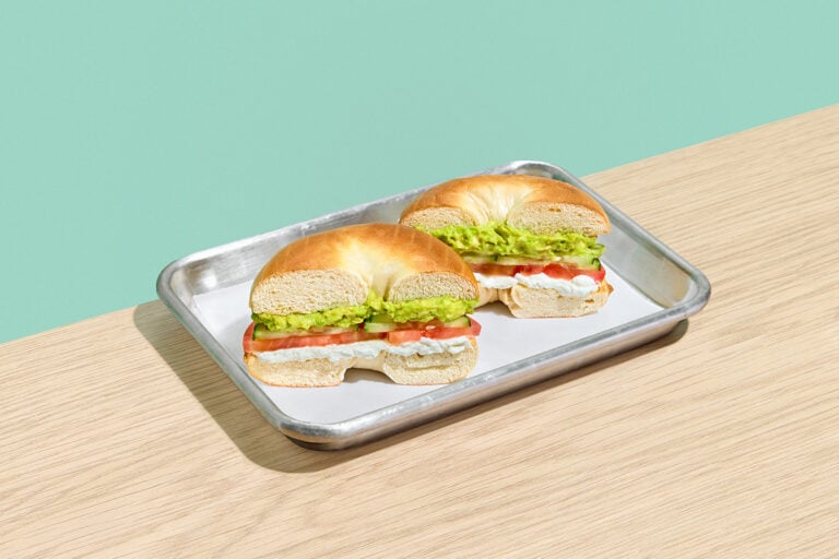 California bagel sandwich with cream cheese, sliced tomato, cucumber, and smashed avocado on a metal tray.