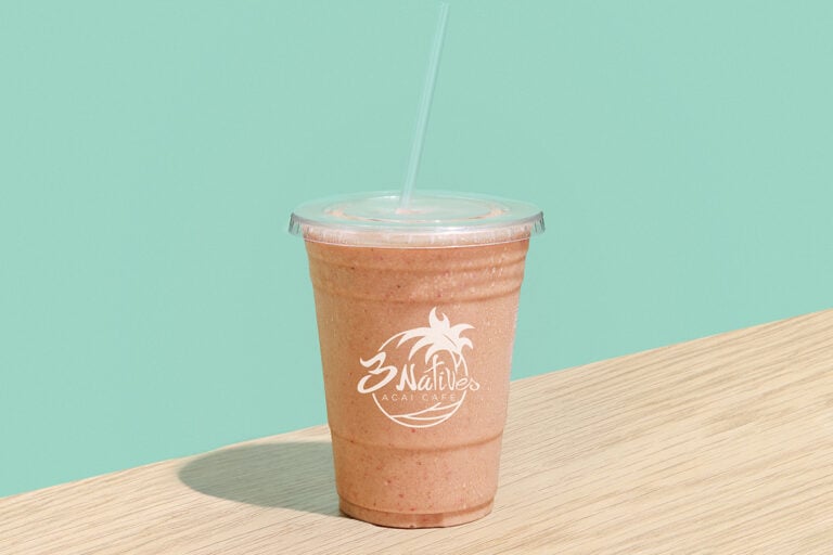 A banana, strawberry, peanut butter, and chocolate protein smoothie made with almond milk is served in a clear plastic cup with a lid and straw on a wooden table against a mint green background. The cup features the “3 Natives Açaí Cafe” logo.