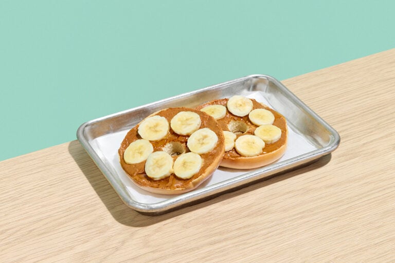 Plain bagel with peanut butter, sliced bananas, and honey on a silver serving tray.