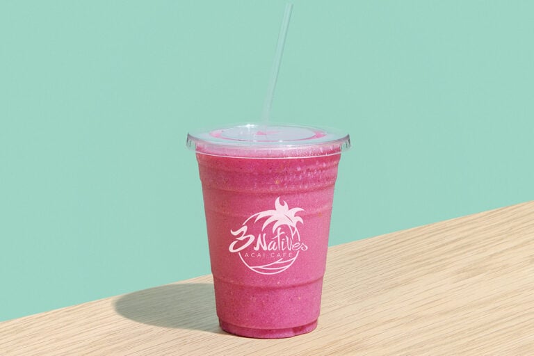 Bright pink fruit smoothie in a clear takeaway cup with straw from 3 Natives Acai Cafe, placed on a wooden table against a pastel mint green background.