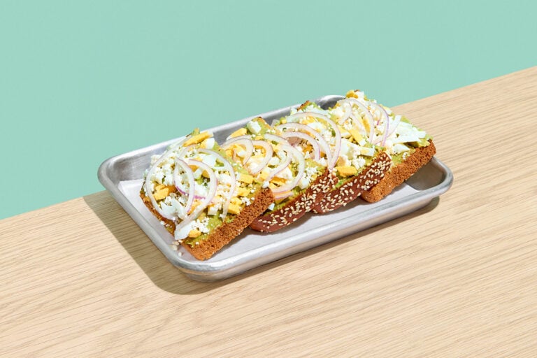 Protein Toast on seeded bread topped with avocado mash, hard-boiled egg, feta cheese, and red onion on a silver tray.