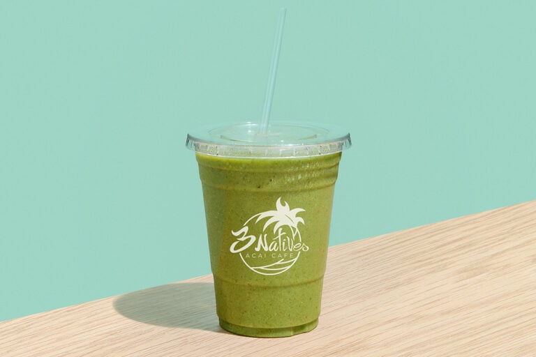 Green smoothie in a clear plastic cup with straw from 3 Natives Acai Cafe, set on a wooden table with a soft mint green background.