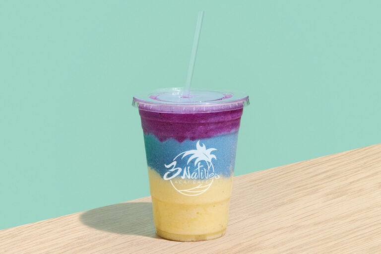 Colorful layered smoothie with yellow, blue, and purple fruit blends in a clear cup from 3 Natives Acai Cafe on a wooden table.